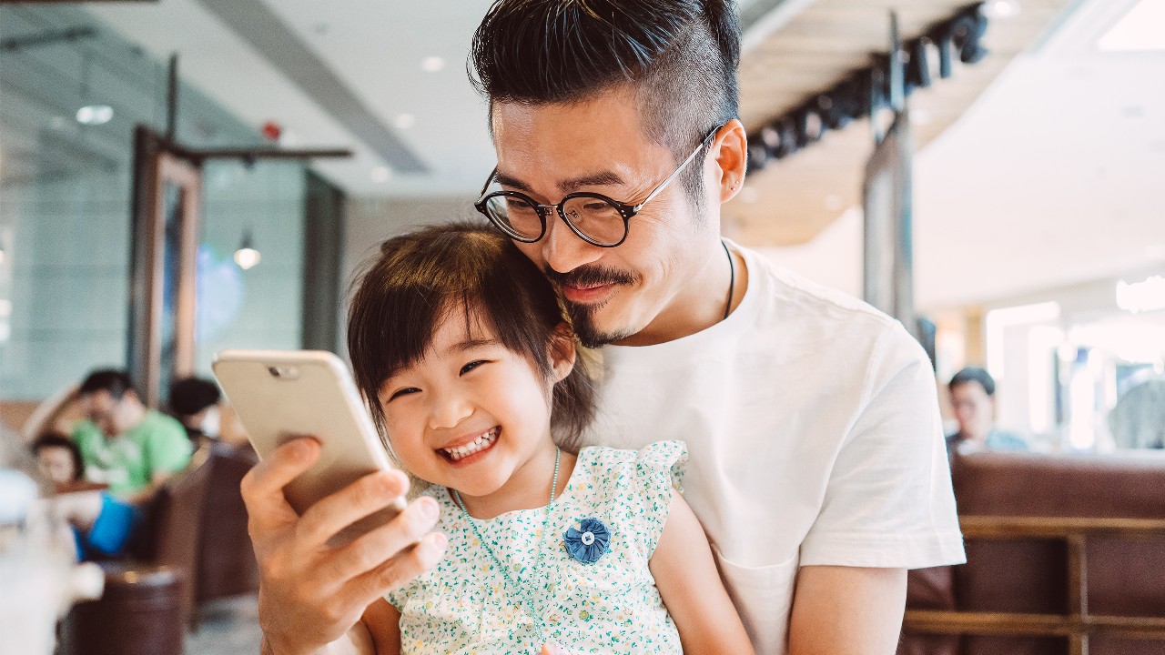 Father and daughter are happily watching mobile together; image used Rack up online spending offers with your HSBC Red Credit Card.