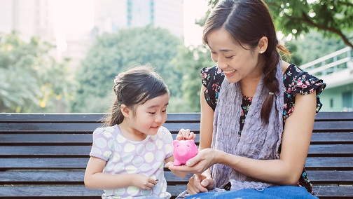 面带笑容的妈妈手托着小猪存钱罐，让女儿投钱币入内；图片使用于汇丰财富管理家庭保障页面。