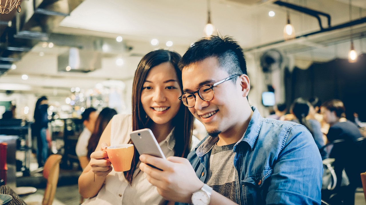 Smiling young woman and man looking at mobile phone while sitting at restaurant; image used for the HSBC Transfer and Payments page.