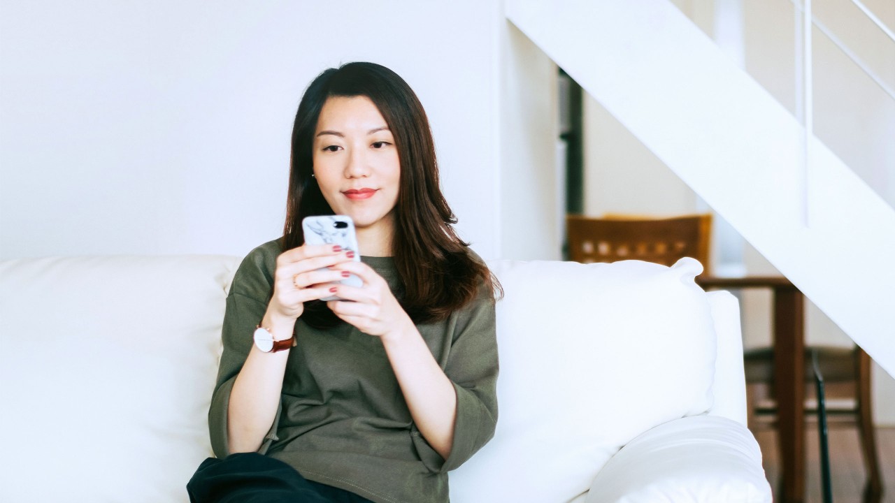 Woman is holding a smartphone on the sofa; image used for "Home loving" article.