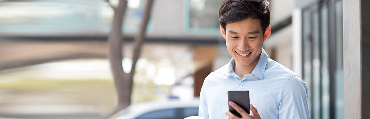 A business man is smiling and holding a smartphone at the street. Image used for HSBC Mobile Branch page.